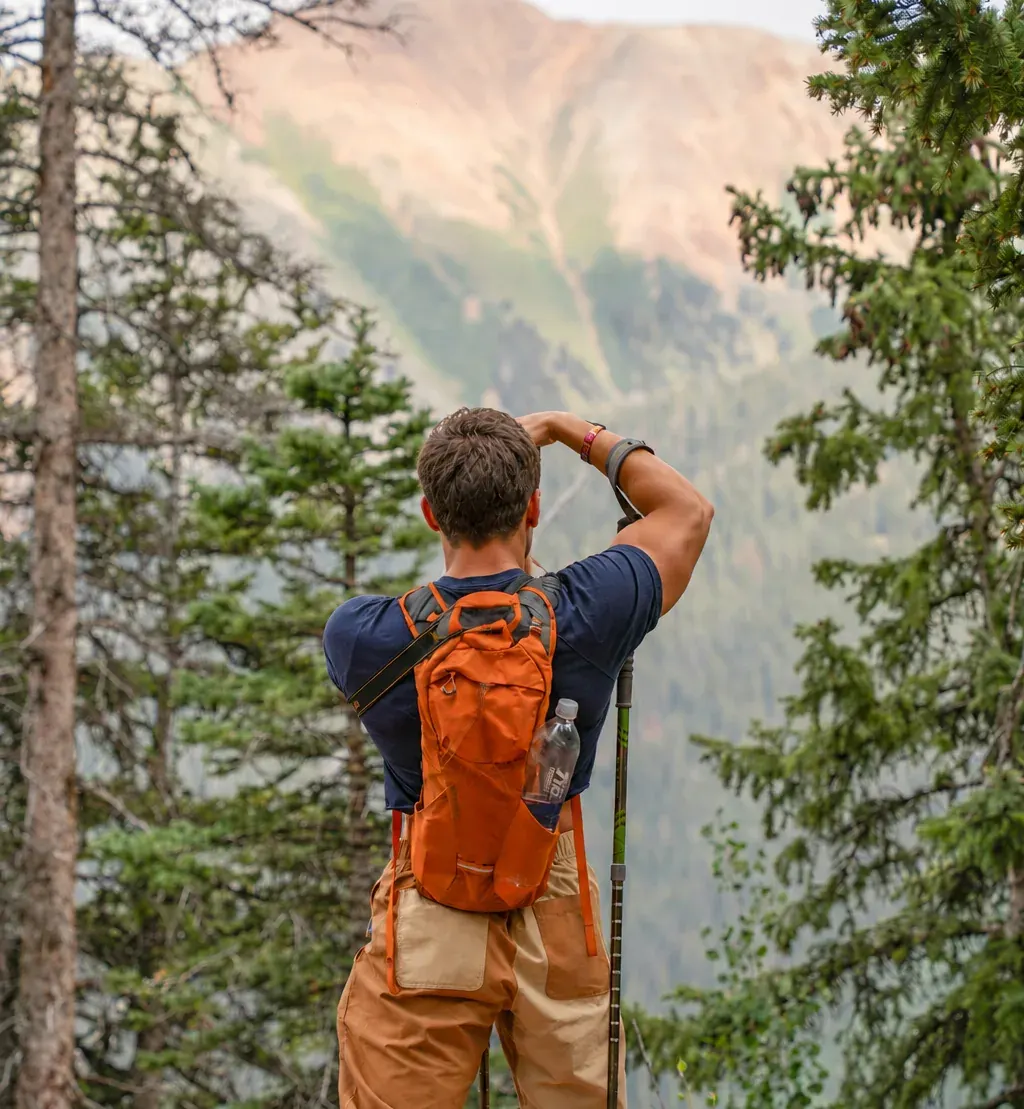 Un senderista con mochila naranja hace una foto a la montaña entre pinos